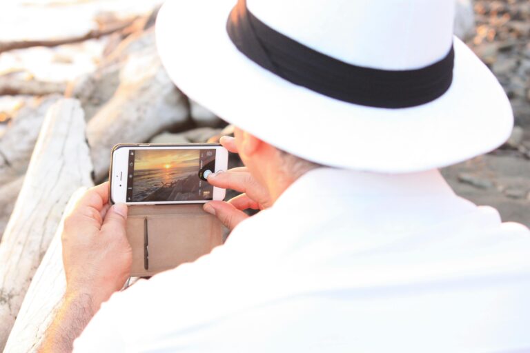A man in a white hat photographs a serene beach sunset using his smartphone.