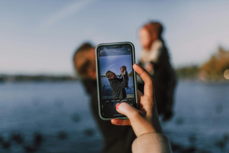 A father lifting his child against a serene outdoor backdrop, capturing the moment on a smartphone.