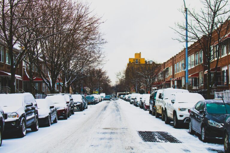 A winter urban scene showing parked cars covered in snow on a residential street lined with bare trees and buildings.