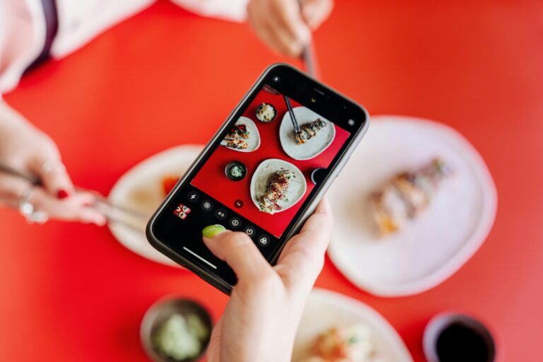 Close-up of hands photographing sushi with a smartphone on a vibrant red table.